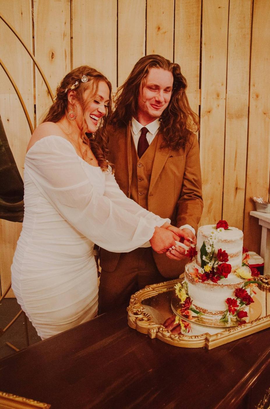A smiling newlywed couple cutting their flower-covered wedding cake