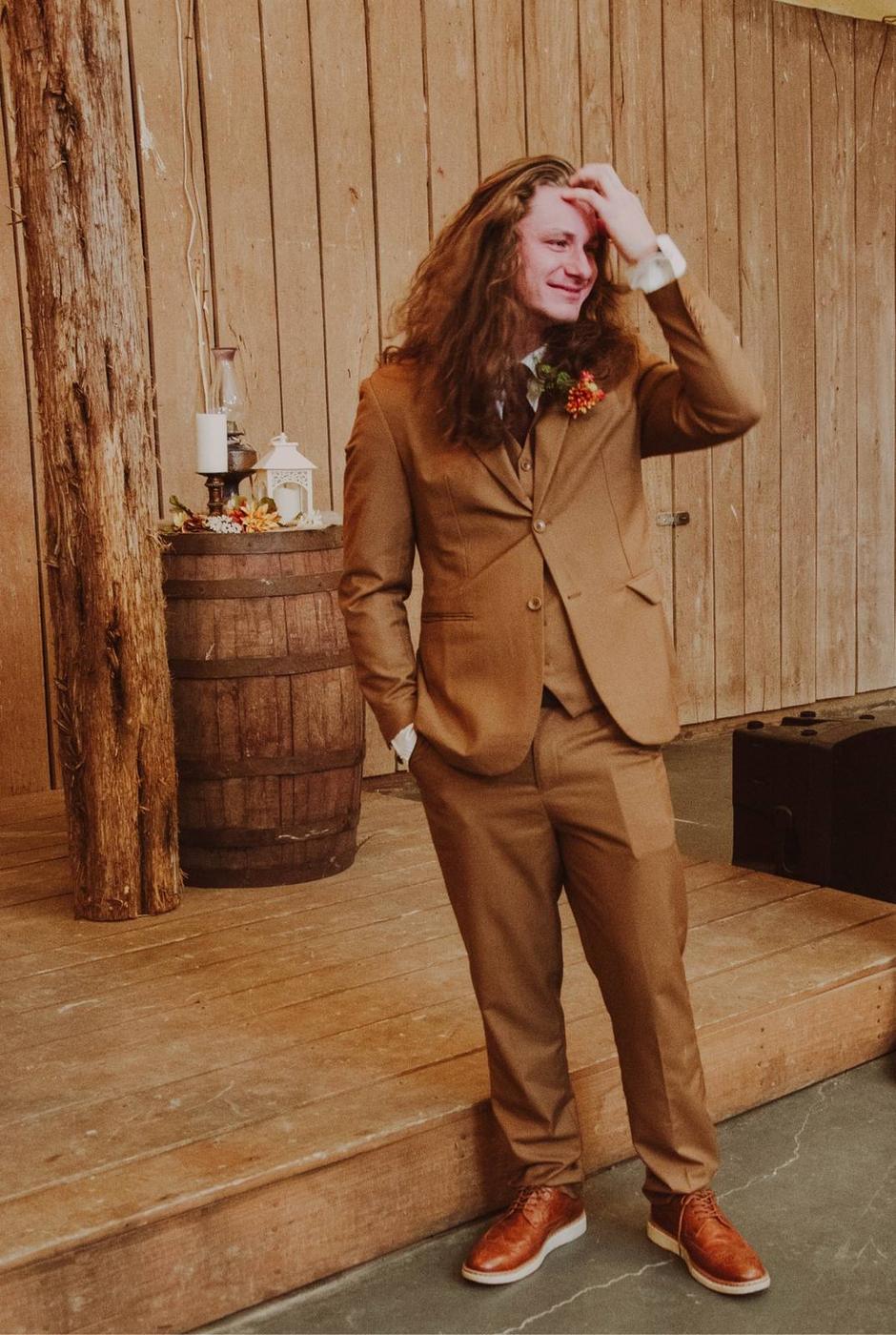 A groom in a brown suit standing before a rustic barn wall