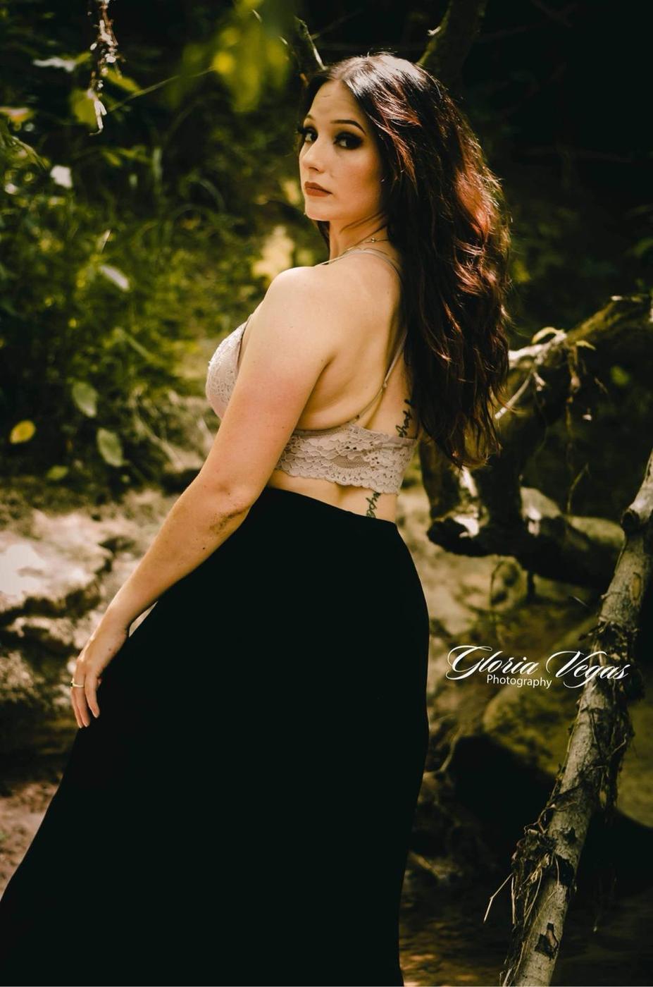 Woman in a lace bralette and long black skirt looking back over her shoulder beside a creek