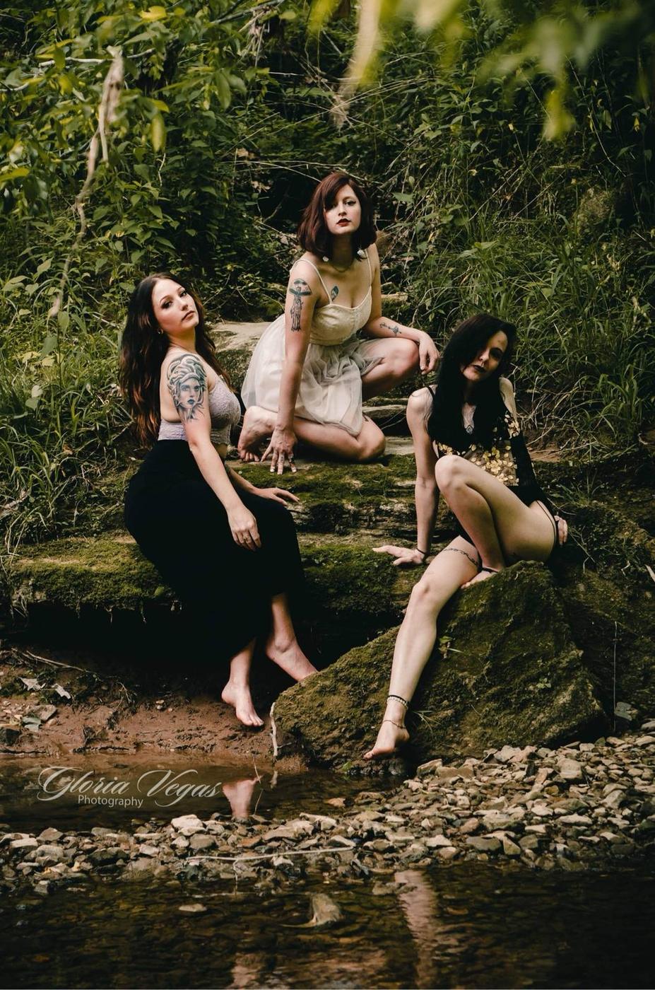 Three women in dresses posing on mossy rocks beside a creek