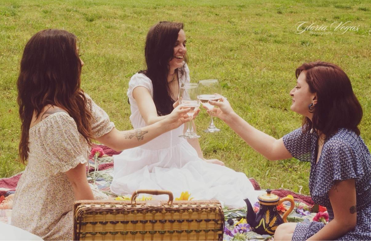 Three women in summer dresses toasting with wine glasses at a picnic